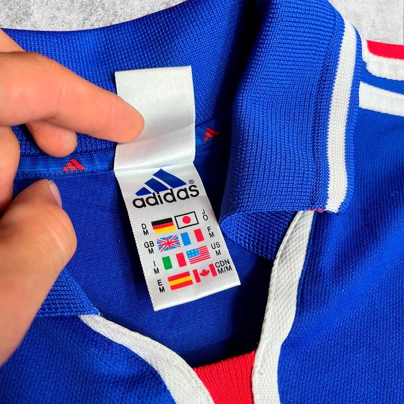 May include: A close-up of a blue Adidas jersey with white stripes and a red accent. The jersey features a white tag with the Adidas logo and country flags. The collar is blue with a white drawstring.