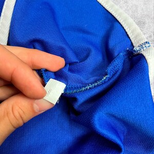 May include: Close-up of a blue athletic garment with white trim. A small white tag with the text "03/00" is held by a hand. The fabric has a textured appearance. The garment appears to be made of a synthetic material.
