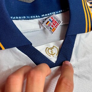 May include: Close-up of a white Real Madrid soccer jersey with navy blue collar and gold accents. The jersey features the Real Madrid crest and Adidas logo. The tag shows international size conversions.