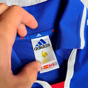 May include: Close-up of a blue Adidas football shirt with white stripes and a red collar. The label shows the Adidas logo, a rooster emblem, and the letters "FFF". The shirt is held by a hand.