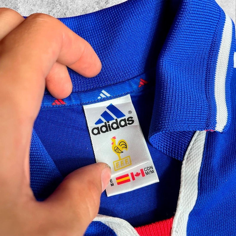 May include: Close-up of a blue Adidas football shirt with white stripes and a red collar. The label shows the Adidas logo, a rooster emblem, and the letters "FFF". The shirt is held by a hand.