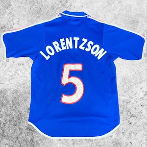 May include: A blue football shirt with the name "LORENTZSON" and the number "5" in white. The shirt has white and red trim on the sleeves and collar. The shirt is made of a breathable material.