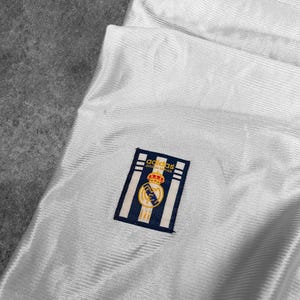 May include: Close-up of a white jersey with a textured pattern. The jersey features a navy blue and gold emblem with the text "adidas OFFICIAL REPLICA". The emblem is on the upper left side of the jersey.