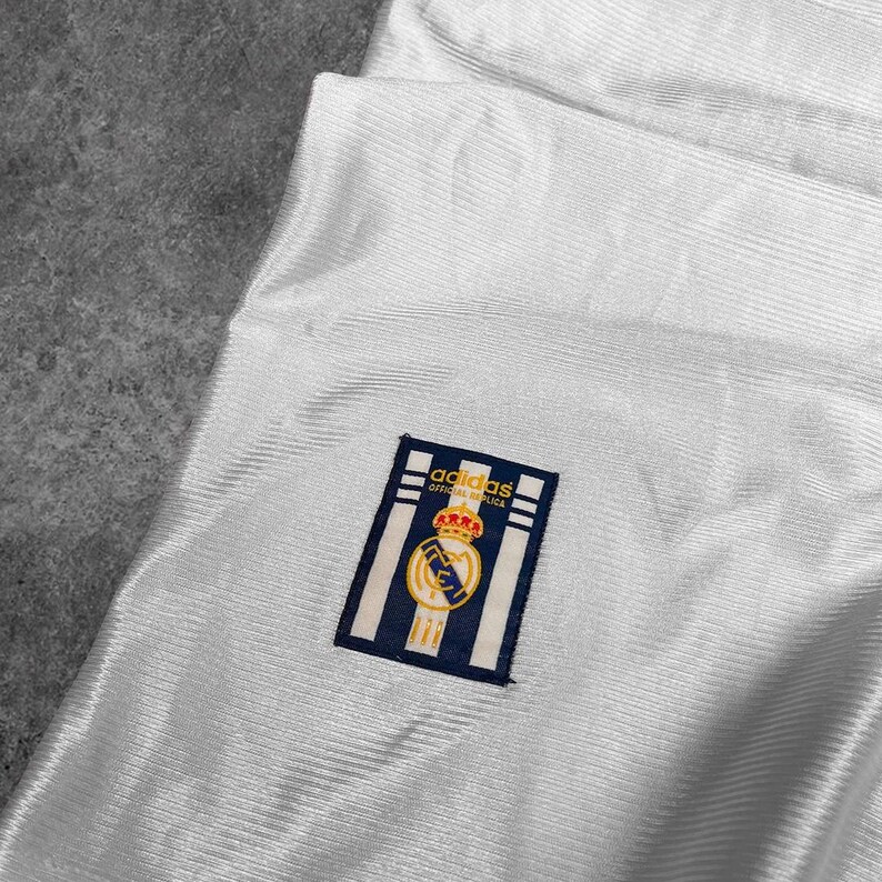 May include: Close-up of a white jersey with a textured pattern. The jersey features a navy blue and gold emblem with the text "adidas OFFICIAL REPLICA". The emblem is on the upper left side of the jersey.