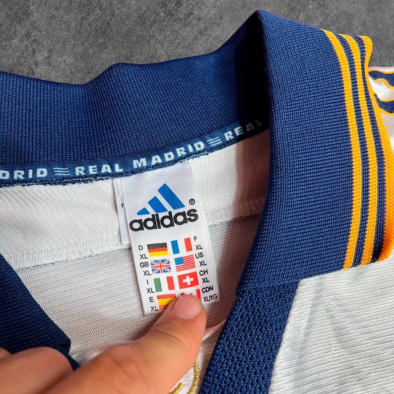 May include: Close-up of a white and navy blue Adidas Real Madrid jersey. The jersey has a navy blue collar and sleeve trim with gold stripes. The Adidas logo and size tag are visible.