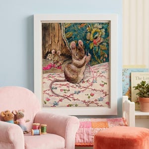 Pode incluir: Ilustração emoldurada de um rato costurando com linha rosa, com outros ratos ao fundo. A obra de arte é ambientada em um fundo com estampa floral. Uma poltrona rosa, bichos de pelúcia de rato e carretéis de linha estão em primeiro plano.