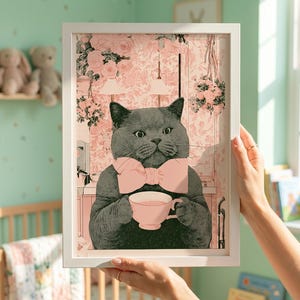 May include: A framed art print featuring a gray cat wearing a pink bow tie and holding a teacup. The background has a pink floral pattern. The print is held by a person in a room with a crib and a shelf with stuffed animals.