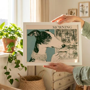 May include: A framed art print featuring a black and white illustration of a dog with the word "MORNING" at the top. The dog is depicted with a cup of tea in a kitchen scene. The print is held up in front of a window.
