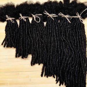 Peut inclure: Six paquets d'extensions de cheveux synthétiques noirs coiffés en fausses dreadlocks. Chaque paquet est attaché en haut avec une fine ficelle en chanvre brun clair. Les paquets sont disposés en escalier, montrant des longueurs variables.