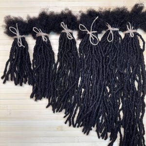 以下が含まれることがあります： カスケード状の斜めのラインに配置された6束の黒い合成ヘアロックス。各束は上部に質感のあるコイル状の外観を持ち、根元の部分で結ばれた細い明るい色の紐で固定されています。