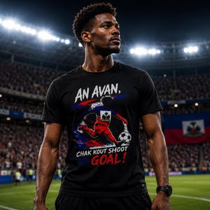 Puede incluir: Camiseta negra con un gráfico de fútbol y el texto "AN AVAN, CHAK KOUT SHOOT GOAL!". La camiseta presenta un jugador de fútbol en rojo y azul, con la bandera haitiana en el fondo. La imagen está tomada en un estadio.