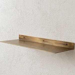 Op de afbeelding: Een wandplank van messingkleurig metaal. De plank heeft een rechthoekige vorm met een plat oppervlak en een verhoogde achterkant, bevestigd aan de muur met schroeven. Het is ontworpen voor het uitstallen van items. Ongeveer 60 cm lang.