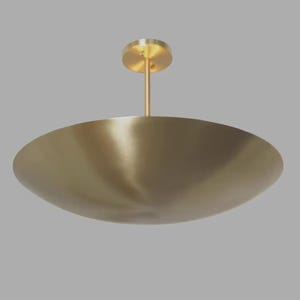 Peut inclure: Un plafonnier moderne de couleur laiton. Le luminaire est composé d'un grand abat-jour peu profond en forme de bol, suspendu à une tige centrale et à un support de plafond circulaire. Design minimaliste.