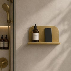 May include: A brass-colored shower shelf holds a brown shampoo bottle and a black smartphone. The shelf is mounted on a light gray tiled wall, with a matching shower head and soap dispensers visible.