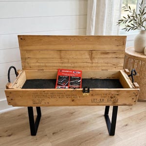 May include: A rectangular wooden storage chest with a hinged lid, black metal U-shaped legs, and black rope handles. The front features stenciled military text including "3315-00-928-4837-C445-1" and "LOADED 9-75". A red Onreva product card rests inside.