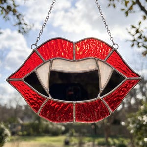 Könnte beinhalten: Ein Buntglas-Suncatcher in Form eines geöffneten Mundes mit spitzen weißen Vampirzähnen. Die Lippen bestehen aus strukturierten roten Glassegmenten, die durch silberne Metalllötlinien verbunden sind. Eine silberne Kette dient zum Aufhängen.