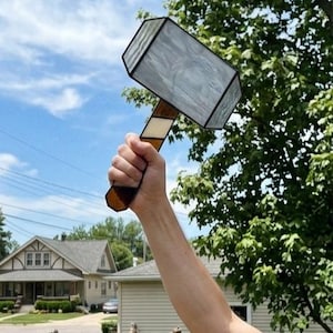 Può includere: Un martello in vetro colorato con una testa rettangolare caratterizzata da pannelli in vetro grigio e bianco marmorizzato incorniciati in metallo nero. Il manico è composto da segmenti alternati di vetro marrone e crema, dal design geometrico.