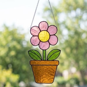 May include: A stained glass suncatcher featuring a pink flower with a yellow center, two green leaves, and a brown pot. The piece is suspended by a thin metal chain, showcasing textured glass panels held together by dark metal solder lines.