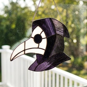 May include: A stained glass suncatcher shaped like a plague doctor mask, featuring deep purple glass for the hood and opaque white glass for the beak and eye area, suspended by a thin metal chain against a window pane.