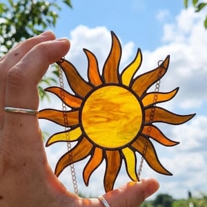 May include: A sun-shaped stained glass suncatcher featuring a central yellow circular piece surrounded by alternating amber and golden yellow flame-like rays, all encased in dark metal solder lines and suspended by a thin silver-toned metal chain.