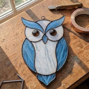 Peut inclure: Un attrape-soleil en vitrail en forme de hibou composé de panneaux de verre texturé blanc et bleu clair avec des yeux en verre brun foncé. Le hibou est bordé d'une soudure métallique sombre et inclut une chaînette de suspension en métal.