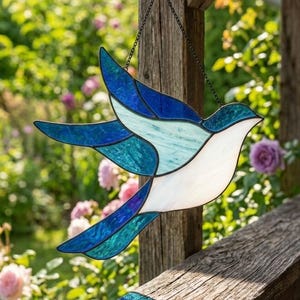 May include: A stained glass suncatcher shaped like a bird in flight, featuring various shades of blue, teal, and white glass pieces joined by dark metal solder lines, suspended by a thin black chain against a blurred garden background.