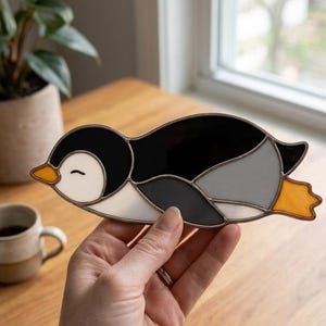Pode incluir: Um apanhador de sol de vitral em forma de pinguim a nadar, com segmentos de vidro preto, branco e cinzento. O pinguim tem um bico e patas cor de laranja brilhante, com um detalhe de olho fechado gravado na face de vidro branco.