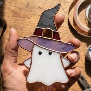 May include: A stained glass ghost ornament featuring a white body with two black circular eyes and a pointed witch hat. The hat is constructed from dark gray and purple glass segments with a copper-colored metal frame holding the pieces together.