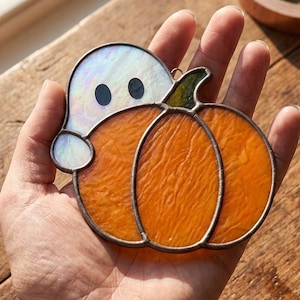 May include: A stained glass suncatcher featuring an iridescent white ghost with two black circular eyes peeking out from behind a textured orange pumpkin. The pieces are joined by dark metal solder lines, creating a decorative seasonal ornament.