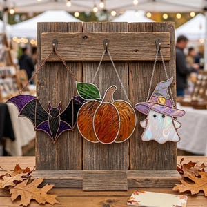 Op de afbeelding: Drie glas-in-lood ornamenten hangen aan een rustiek houten displaybord. De ornamenten bestaan uit een zwarte en paarse vleermuis, een oranje pompoen met een groen blad, en een witte geest met een paarse en gele heksenhoed en zwarte ogen.