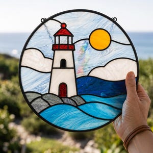 May include: A circular stained glass suncatcher featuring a white lighthouse with a red roof, a bright yellow sun, white clouds, and blue ocean waves. The piece is framed in black metal and depicts a coastal scene with rocky terrain.