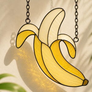 May include: A stained glass banana suncatcher featuring segments of pale yellow and vibrant golden yellow glass held together by dark metal solder lines. The piece is suspended by two black metal chains attached to the top of the banana shape.