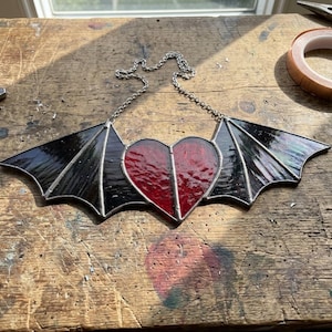 May include: A stained glass suncatcher featuring a central red heart flanked by two black bat wings. The glass pieces are joined with dark metal solder lines, and a silver-toned metal chain is attached to the top for hanging purposes.