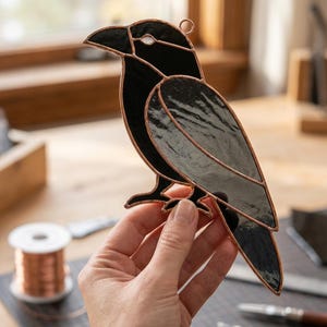 May include: A stained glass raven suncatcher featuring opaque black glass pieces joined by copper foil solder lines. The bird is depicted in profile with a small circular hanging loop at the top, showcasing a clean, segmented design.