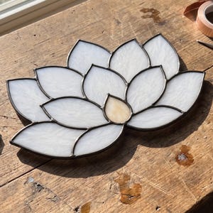 May include: A stained glass lotus flower ornament featuring multiple white, textured glass petals arranged in a symmetrical bloom pattern. The centre petal is a pale, translucent amber colour. Each individual glass piece is encased in dark metal solder lines.