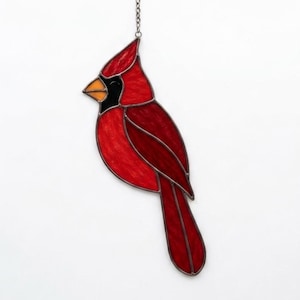 May include: A stained glass suncatcher shaped like a cardinal bird, featuring vibrant red glass panels, a black face mask around the eye, and a yellow beak. The bird is outlined in dark metal solder and hangs from a small silver-coloured metal chain.