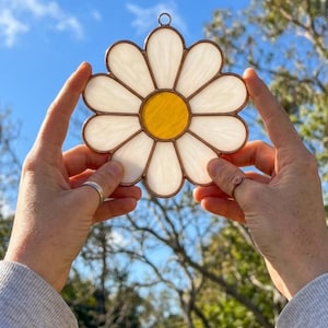 Puede incluir: Un atrapasoles de vidrieras con forma de flor de margarita con doce pétalos blancos opalescentes que rodean un centro circular de color amarillo vibrante. Las piezas de vidrio individuales están unidas por finas líneas de soldadura de metal oscuro.
