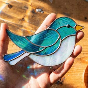 May include: A stained glass bird suncatcher featuring a vibrant teal and blue wing and back, a white iridescent breast, and a small yellow beak. The glass pieces are joined by dark metal solder lines, and a small metal loop is attached to the top.