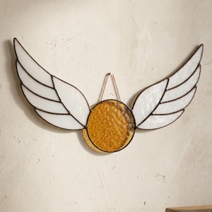 Op de afbeelding: Een glas-in-lood wandhanger met een centraal rond stuk amberkleurig getextureerd glas, geflankeerd door twee witte glazen vleugels. De onderdelen zijn verbonden door donkere metalen soldeerlijnen en er is een dunne metalen ketting voor ophanging.