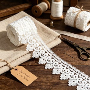 Puede incluir: Un rollo de encaje blanco con borde festoneado que presenta intrincados patrones florales y de remolinos. El encaje se coloca sobre una tela beige doblada junto a una etiqueta de papel marrón con la inscripción Handmade Lace Trim, tijeras y carretes.