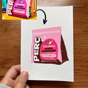 May include: A white print featuring a stylized illustration of a pink coffee bag with the text PERC, COLOMBIA YOUNG PRODUCERS, Pink Starburst & Frosting, and a flavor intensity scale. The bag has a dark brown side panel and a pink label with icons.