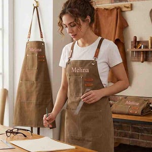 May include: A brown canvas apron featuring adjustable leather straps, metal hardware, and multiple pockets. The name Melina is embroidered in light-coloured thread on the chest. The design includes a large lower pocket and a smaller upper pocket.