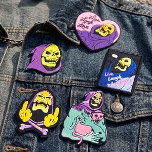 May include: Five embroidered patches featuring Skeletor characters attached to a blue denim jacket. One heart-shaped patch reads Live Laugh Love. A rectangular patch also reads Live Laugh Love. Other patches show various expressions and poses.