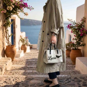 May include: A beige trench coat with black buttons and a white handbag featuring black handles, the name Lucia in black script, and a small black whale graphic. The handbag is held by a hand wearing a silver watch and rings.