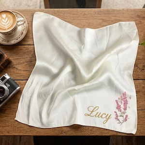 Pode incluir: Lenço de seda marfim com o nome "Lucy" bordado em ouro, acompanhado por um desenho floral rosa e verde. O lenço está sobre uma superfície de madeira, junto a uma câmera vintage e uma xícara de café.