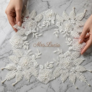May include: A white lace appliqué featuring intricate floral and leaf embroidery with small pearl accents. The centre of the sheer fabric displays the text Mrs. Lucia in a cursive font. The edges are adorned with dangling pearl beads.