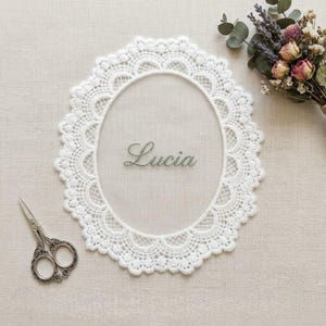 Könnte beinhalten: Ein ovales weißes Spitzendeckchen mit gewelltem Rand und einem komplexen Netzmuster. Der Name Lucia ist mittig in grüner Schreibschrift eingestickt. Eine kleine Schere aus silberfarbenem Metall befindet sich unten links im Bild.