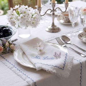 May include: A white linen napkin with frayed edges, embroidered with the blue text Ms. Lucia and a small lavender sprig. The napkin rests on a white plate atop a matching tablecloth with blue border stitching, near a silver candelabra and flowers.