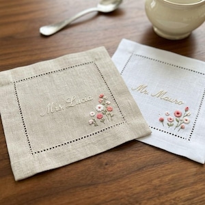 Puede incluir: Dos servilletas cuadradas de lino con bordes de vainica. Una es beige con el texto bordado Mrs. Lucia y un pequeño motivo floral. La otra es blanca con el texto bordado Mr. Mauro y un diseño floral a juego en tonos rosas y blancos.