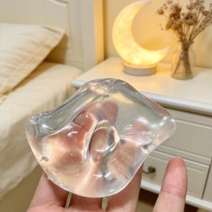 Könnte beinhalten: Eine klare, halbkugelförmige Brustprothese aus Silikon mit einer glatten, gelartigen Textur. Das Objekt ist transparent, sodass die Haut darunter sichtbar ist, und weist eine abgerundete, konturierte Form auf, die der Anatomie nachempfunden ist.
